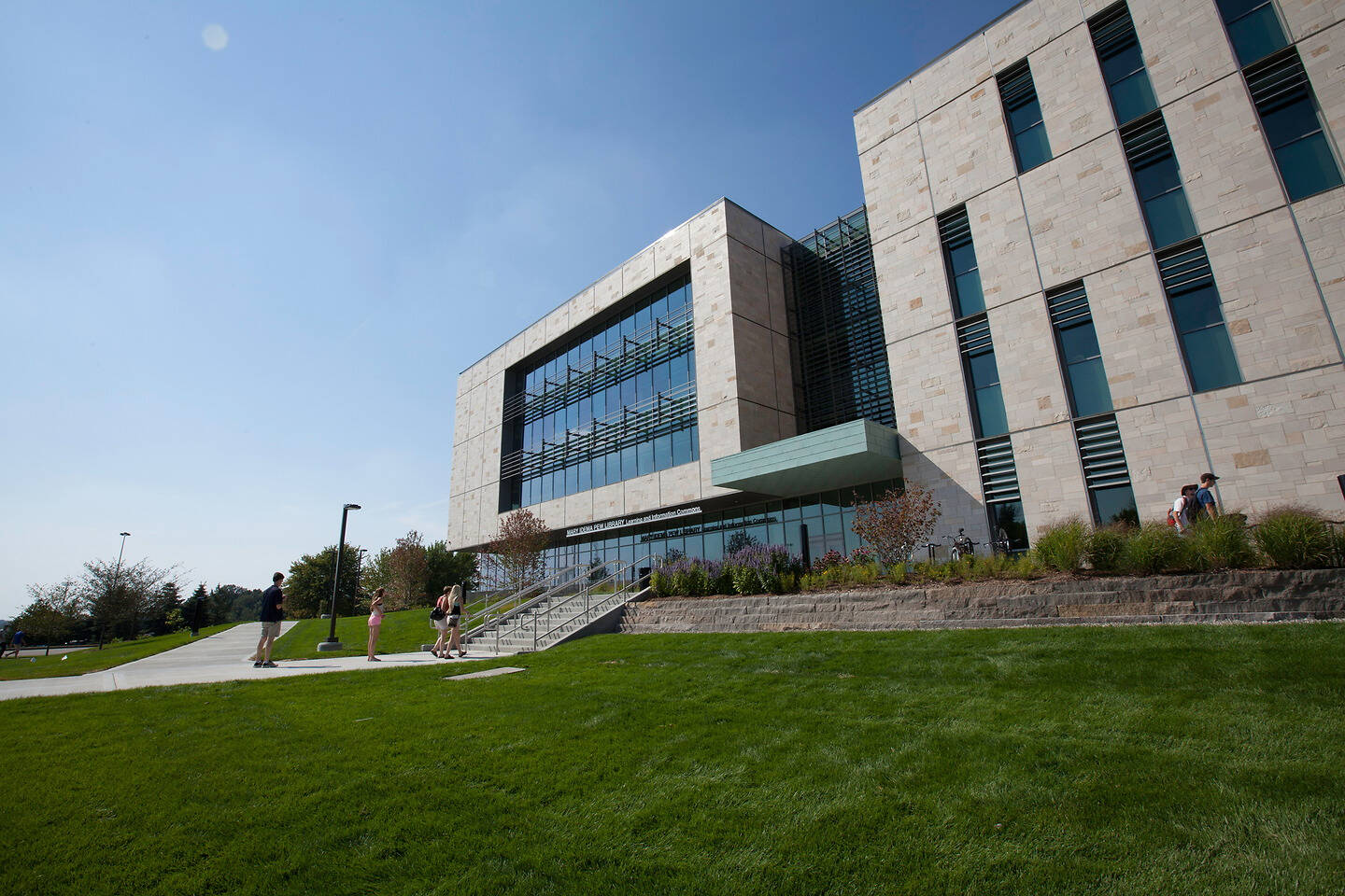 A modern building with stone and glass facades stands under a clear blue sky, with people walking on a grassy lawn and sidewalk nearby.